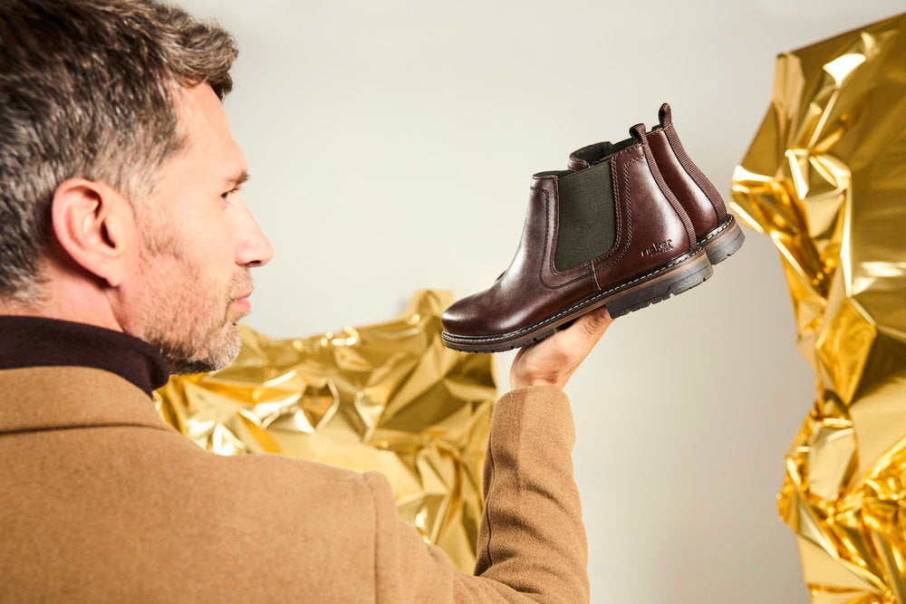 man wearing beige jacket holding a pair of brown leather mens chelsea boots by Rieker with gold back drop