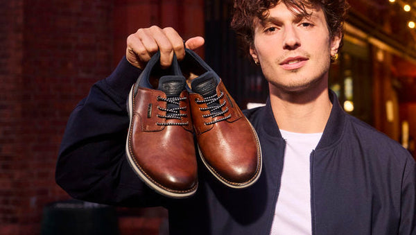 A man holding up a pair of brown leather lace-up dress shoes, wearing a dark navy bomber jacket over a white t-shirt, standing in a dimly lit urban alleyway at night with warm string lights in the background.